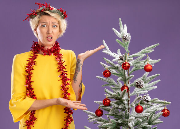 立场笨手笨脚的金发女郎戴着圣诞花环 脖子上戴着金属丝花环 站在装饰好的圣诞树旁 用手指着圣诞树 在紫色的墙上显得孤立无援脖子头花环