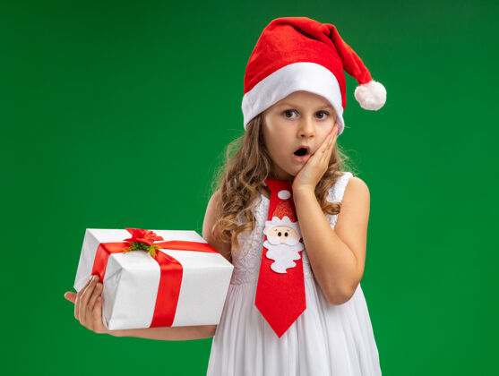 抱害怕的小女孩戴着圣诞帽打着领带拿着礼盒把手放在脸颊上隔离在绿色的墙上女孩圣诞节害怕