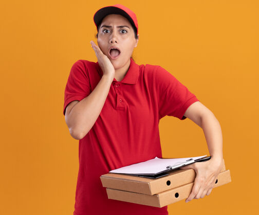 盒子害怕的年轻送货女孩穿着制服 戴着帽子 拿着比萨饼盒和剪贴板 把手放在隔离在橙色墙上的脸颊上人送货站着