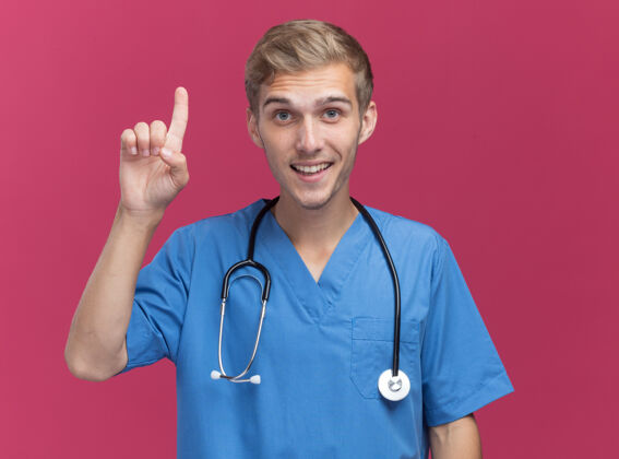 立场印象深刻的年轻男医生穿着医生制服用听诊器点在粉红色的墙上孤立起来感情人医生