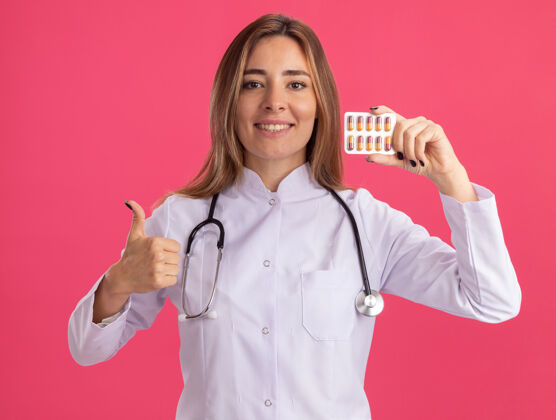衣服面带微笑的年轻女医生穿着医用长袍 手持听诊器的药片 在粉红色的墙上孤立地竖起大拇指微笑人长袍