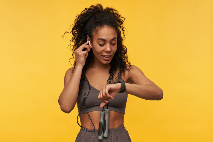 运动快乐迷人的年轻女运动员脖子上跳绳使用智能手表和听音乐使用无线耳机隔离黄墙无线手表肖像