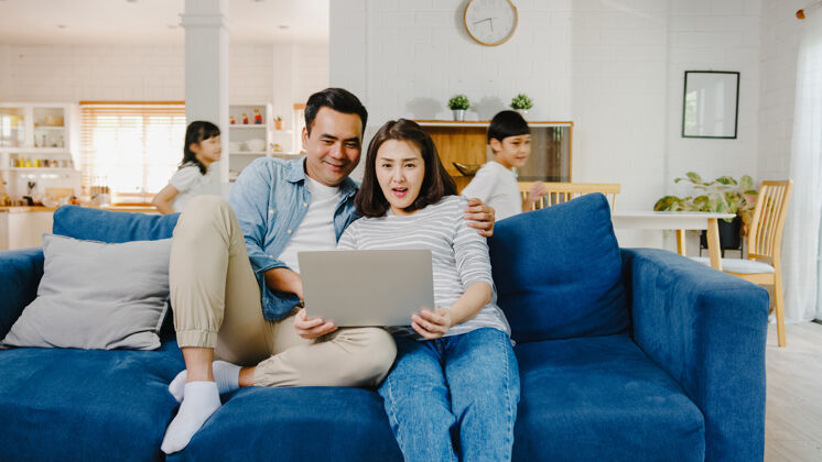 男人亚洲家庭的爸爸妈妈坐在沙发上享受着用笔记本电脑上网购物的乐趣 而女儿和儿子则在家里的客厅里围着沙发大喊大叫男孩家庭女儿