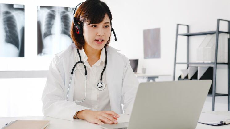 日语年轻的亚洲女医生 身穿白色医疗制服 手持听诊器 使用笔记本电脑 在诊所或医院的办公桌上与患者进行视频会议视频亚洲人帮助