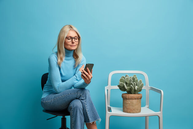 水平女人戴着透明眼镜整洁的衣服在网上看新闻拿着手机排队在椅子上摆姿势孤独地躺在蓝色的椅子上高级皱纹女士