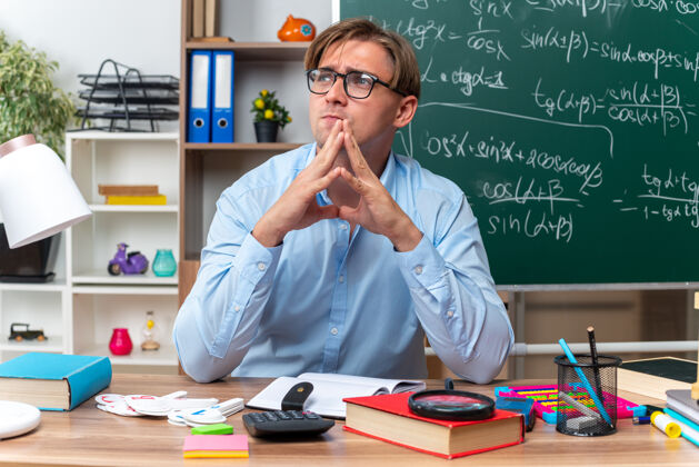佩戴年轻的男老师戴着眼镜坐在课桌旁 手里拿着书和笔记 双手合十 脸上带着沉思的表情 在教室的黑板前思考黑板手掌表情