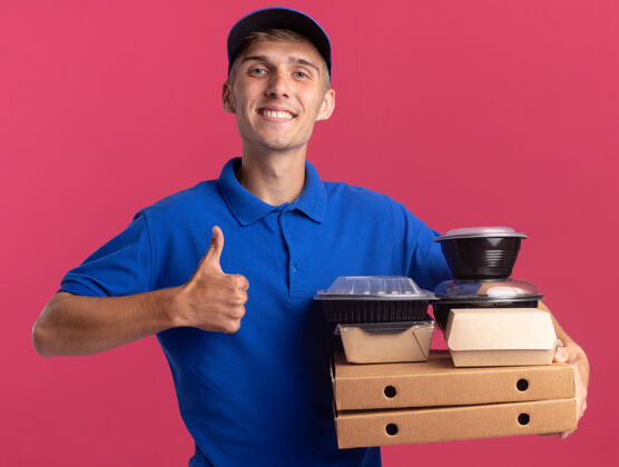 比萨饼微笑着的年轻金发送货员竖起大拇指 拿着比萨饼盒上的食品容器和包装容器粉红色男孩