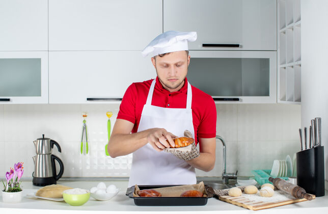 持有人白色厨房里 年轻忙碌的男厨师手持一个新鲜出炉的糕点 戴着支架 正对着眼前的景象帽子观点男人
