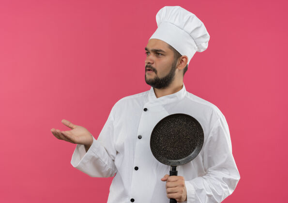 年轻困惑的年轻男厨师身着厨师制服 手里拿着煎锅 空手看着隔离在粉红色墙上的一面制服厨师手