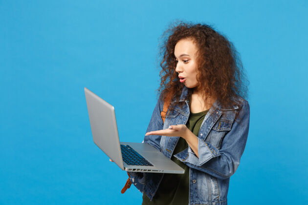女孩年轻的非洲裔美国女孩十几岁的学生在牛仔服装 背包工作的pc隔离在蓝色的墙壁上设备包电脑