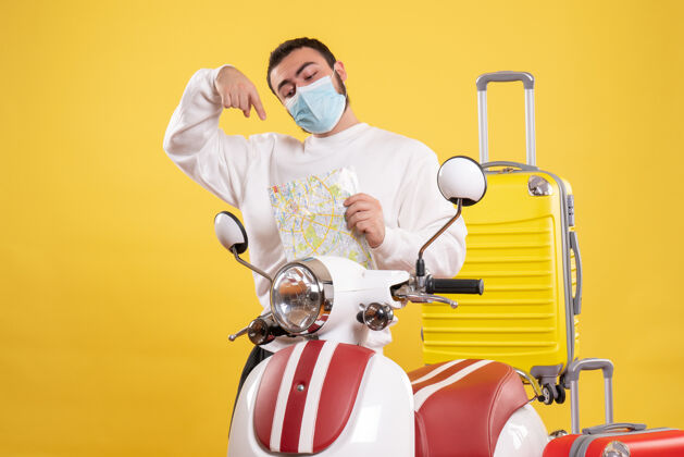 顶部旅行概念的俯视图 戴着医用面罩的自信的家伙站在摩托车旁边 黄色手提箱在上面 指着地图地图黄色运动