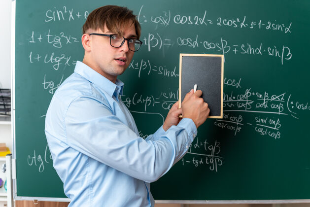黑板戴着眼镜拿着小黑板和粉笔的年轻男老师惊讶地站在教室里数学公式的黑板旁边教室年轻数学