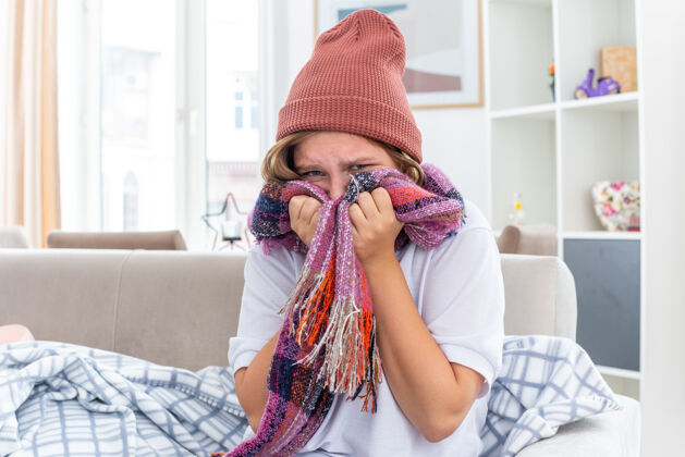 温暖不健康的年轻女子戴着暖和的帽子 脖子上围着围巾 感觉不舒服 生病 患感冒和流感 看起来很担心 坐在客厅里的沙发上周围年轻担心