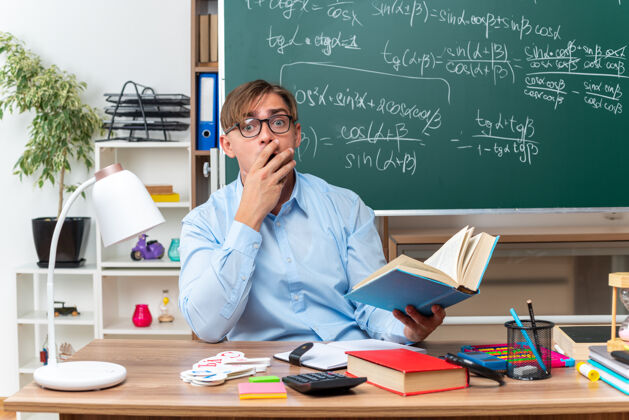 穿年轻的男老师戴着眼镜拿着书被震得捂着嘴坐在教室黑板前的书桌旁拿着书和笔记学校教室年轻