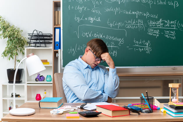 困惑戴着眼镜的年轻男老师坐在教室的黑板前 手里拿着书和笔记 神情迷茫 非常焦虑学校教室焦虑