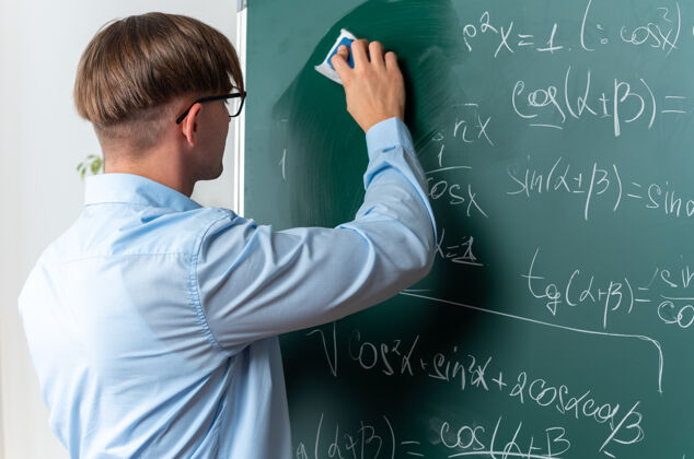 站自信的年轻男老师戴着眼镜站在教室黑板边擦数学公式黑板近擦