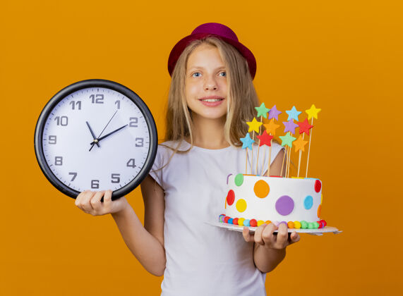 时钟戴节日帽的漂亮小女孩拿着生日蛋糕和挂钟 笑脸盈盈 生日派对概念微笑蛋糕站