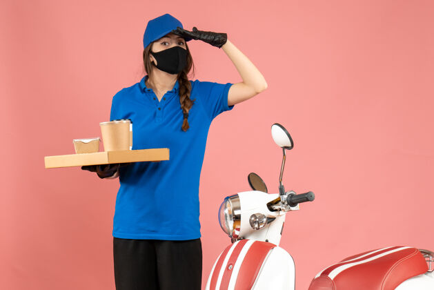 摩托车前视图快递女孩戴着医用口罩手套站在摩托车旁拿着咖啡小蛋糕专注于粉彩桃色背景上的东西穿焦点人