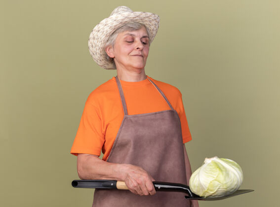 卷心菜自信的上了年纪的女园丁戴着园艺帽 手里拿着铲子上的白菜穿举行花园