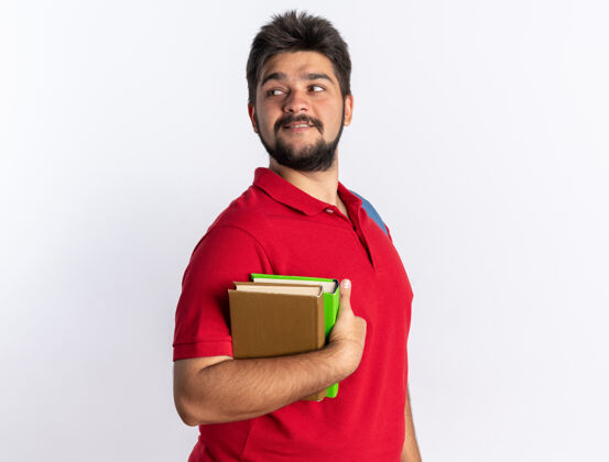 姿势年轻的留着胡子的学生 穿着红色马球衫 背着书包 拿着笔记本 面带微笑 自信地站在白色的墙上背包人年轻人
