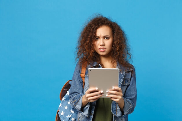 打字年轻的非洲裔美国女孩十几岁的学生在牛仔服装 背包举行垫电脑隔离在蓝色的墙壁上显示器民族女人