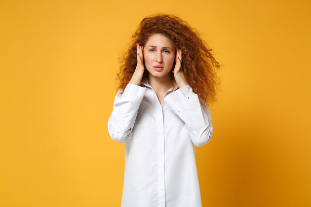 耳朵疲惫的年轻红发女子穿着休闲白衬衫在黄橙色的墙上摆着孤立的姿势人成人一个