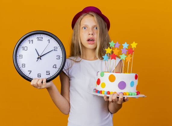 橙色漂亮的小女孩戴着节日帽子拿着生日蛋糕和挂钟惊讶地说 生日派对的概念站生日节日