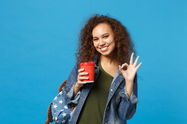 红色年轻的非洲裔美国女孩十几岁的学生在牛仔服装 背包举行纸杯隔离在蓝色的墙壁上能量咖啡马克杯
