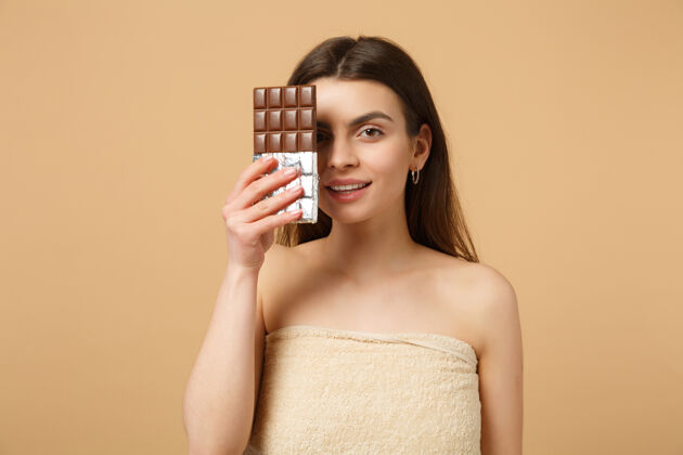 深色近距离拍摄完美肌肤的半裸女人 裸妆将巧克力棒隔离在米色粉彩墙上脸甜点身体护理