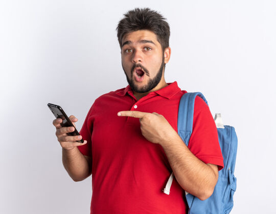移动年轻的留着胡子的学生 穿着红色马球衫 背着背包 拿着智能手机 用食指指着它 站在白墙上 看上去很惊讶姿势马球年轻