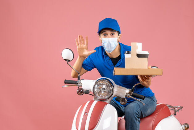 男戴着帽子戴着面具坐在滑板车上递送订单的男性送货员的正视图 在柔和的桃色背景上显示十个桃子面具坐着
