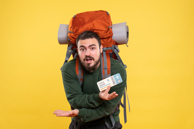 肖像年轻迷茫的旅行小伙背着背包 在黄色背景下出示车票的正面图票显示背包