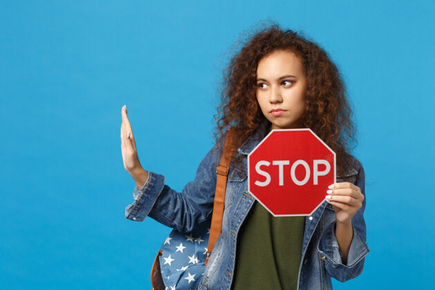 民族年轻的非洲裔美国女孩十几岁的学生在牛仔服装 背包举行停止孤立的蓝色墙壁红色警告手