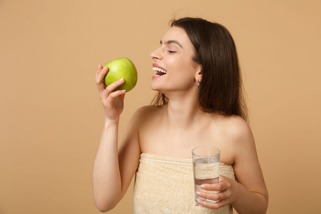 美丽近距离拍摄完美肌肤的半裸女人 裸妆将苹果和水隔离在米色粉彩墙上素食主义者液体脸