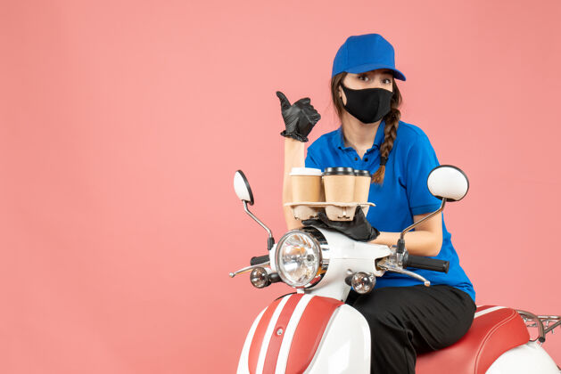 摩托车前视图好奇的女性送货员戴着医用口罩和手套坐在滑板车上拿着订单指着粉彩桃背景前面滑板车女性