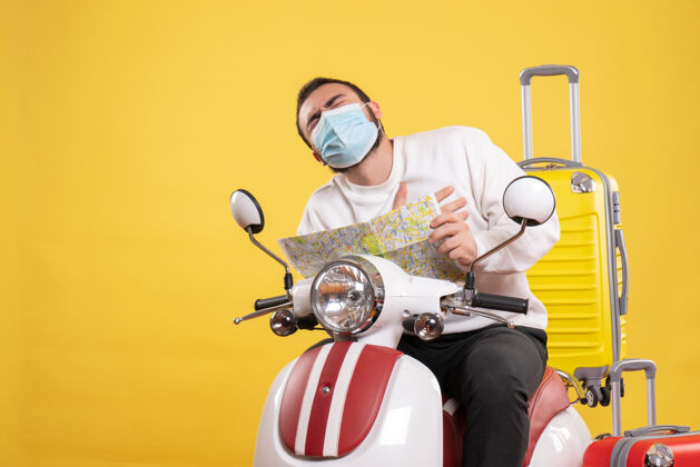 男人旅行理念：戴着医用面罩的年轻人坐在摩托车上 背着黄色手提箱 手里拿着心脏病发的地图攻击坐着黄色