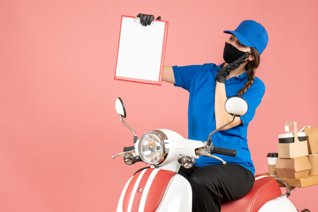 背景顶视图好奇的女快递员戴着医用口罩和手套坐在踏板车上 拿着空纸 在粉彩桃色背景下传递订单举行坐桃