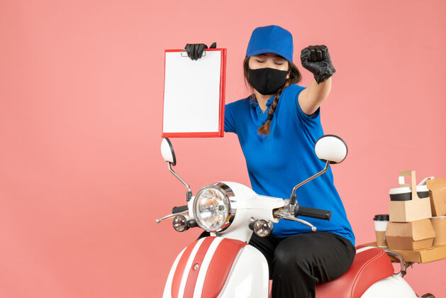 纸俯视图：戴着医用口罩和手套的快乐女快递员坐在滑板车上 手里拿着空纸 在柔和的桃色背景下传递订单摩托车粉彩持有