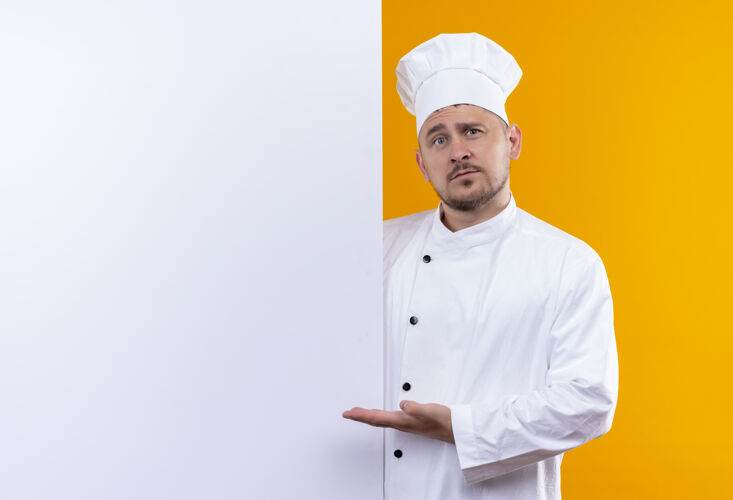 年轻穿着厨师制服的年轻帅哥站在白色的墙后 指着隔离在橙色墙上的复制空间墙壁站立帅气