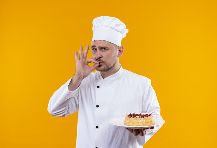 美味自信的年轻帅哥厨师身着厨师制服 端着一盘蛋糕 做着美味的手势孤立在橙色的墙上拿着帅气自信