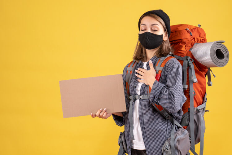 面罩带着黑色面具和背包的旅行女孩把手放在胸前拿着纸板防护箱子硬纸板