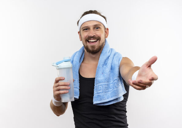 运动快乐的年轻英俊的运动男子戴着头带和腕带拿着水瓶 伸出手隔离在白色的墙上佩戴欢乐瓶子