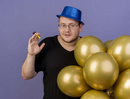 举行高兴的成年斯拉夫人戴着眼镜 戴着蓝色的派对帽 手持氦气球 手持派对哨子成人请帽子