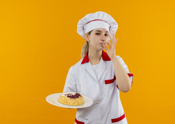 厨师自信的年轻漂亮厨师 身着厨师制服 拿着一盘蛋糕 做着美味的手势 被隔离在橙色的墙上蛋糕年轻自信