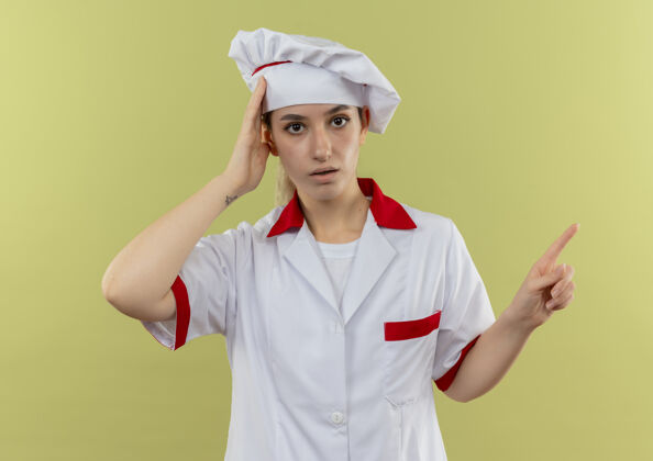 头年轻漂亮的厨师穿着厨师制服 手放在头上 指着隔离在绿色墙上的一侧漂亮印象年轻