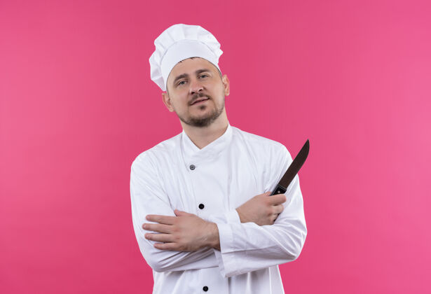 刀自信的年轻帅哥厨师身着厨师制服 站着封闭的姿势 拿着隔离在粉红色墙上的刀帅气封闭厨艺