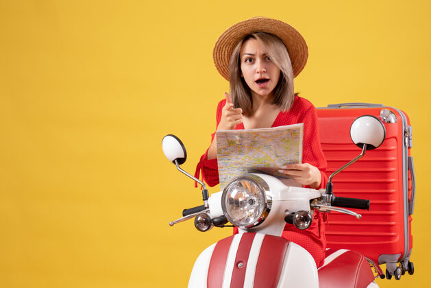 地图骑着轻便摩托车的漂亮女孩拿着红色的手提箱拿着地图指着摄像机帽子人红色