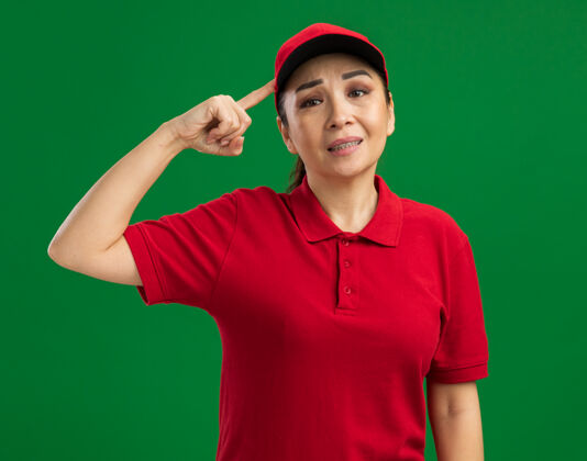 制服身穿红色制服 头戴鸭舌帽的年轻女送货员困惑不解 不高兴地用食指指着站在绿墙上的头指向年轻站着