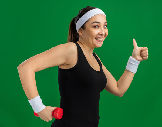 健身头戴哑铃的年轻健身女士微笑着竖起大拇指站在绿色的墙上做运动表演头带女人