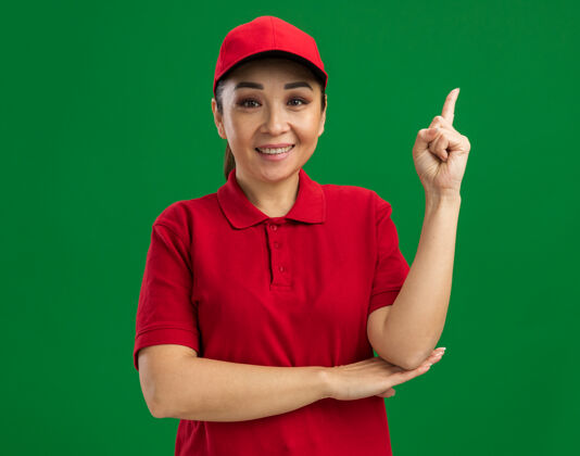 女人身着红色制服 头戴鸭舌帽的年轻女送货员快乐而积极地伸出食指微笑自信地站在绿色的墙上帽子微笑交货